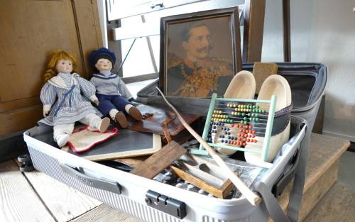 Das Foto eines geöffneten Koffers, in dem sich Holzschuhe, zwei Puppen, ein Portraitbild von Kaiser Wilhelm II, ein Schwamm, ein Rechenschieber eine Kreidetafel und Schwarz-weiß Fotografien befinden.